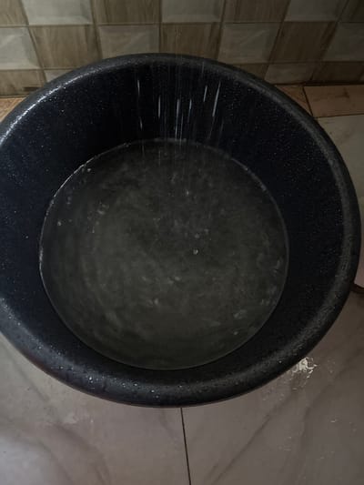 An overhead shot depicts a dark-colored, round plastic bucket containing water, positioned on a tiled floor. The bucket occupies the majority of the frame, with its interior filled with murky water. Streaks of white, indicating splashing or pouring water, are visible on the left inner rim of the bucket and extending into the water.

The floor surrounding the bucket is covered with square tiles, appearing in shades of brown and beige. Some areas of the tiled floor, particularly near the right side of the bucket, show wetness or water splashes. The texture of the bucket's outer surface appears slightly rough and is dotted with small water droplets. The water inside the bucket has a somewhat cloudy or opaque appearance, with faint reflections or light patches on its surface.

The lighting is somewhat dim, suggesting an indoor setting. There are no people visible, and no discernible text or branding on the bucket or the surrounding environment. The scene captures an ordinary moment of water collection or use within a domestic context in Akodo, Nigeria.