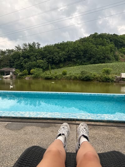 A vertical, eye-level shot depicts a landscape next to a pool, with the photographer’s legs and silver sneakers appearing in the foreground, situated on what looks like a patterned outdoor seating area.
Beside the pool is a murky body of water, possibly a lake or river, with a reflection of the surrounding trees and sky visible on its surface. There is also a small structure, possibly a boathouse, seen on the bank. Two white waterfowl float in the lake.
Across the body of water, a dense forest rises up a hillside, with power lines visible in the cloudy sky above. Based on the prompt, the location is identified as being in the city of Jinhua, China.