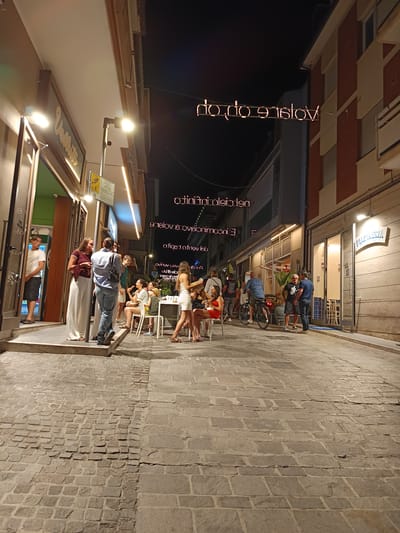 This is a night-time, eye-level, wide shot of a street scene in Roseto degli Abruzzi, Italy, with several people gathered outside establishments. The cobblestone street occupies the foreground, with a slight shift in paving material towards the right. On the left, an open doorway reveals a dimly lit interior with a green wall. A man in a white t-shirt and dark shorts stands just inside the doorway. To his right, a group of people are conversing on a raised platform. Further down the street, on the right side, a building with red and beige sections has windows with closed shutters. A sign with illuminated lettering that reads "PIZZERIA" is visible above a storefront.

In the mid-ground, several people are seated at tables and standing around in small groups. Some are eating or drinking. A woman in a short white dress is walking past a table with chairs. Two men are talking near a bicycle. Overhead, illuminated neon signs form phrases in Italian, with one prominently displaying "VOLARE OPTION". The atmosphere appears social and relaxed, suggesting a lively evening in a commercial area.