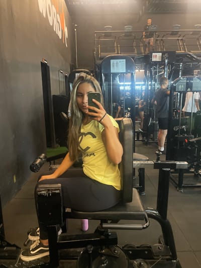 This image captures the interior of a gym in Anápolis, Brazil, as seen through a mirror selfie.

In the foreground, a young adult female is seated on a black padded inner/outer thigh abduction machine. She has long, light brown/blonde hair and is wearing a bright yellow short-sleeved top with a stylized black logo featuring a prominent 'C' or 'G' and the text 'UMA' beneath it. She wears black leggings and black Vans Old Skool sneakers with white laces and soles. Her right hand holds a dark-colored smartphone, likely an iPhone, which she is using to take the photograph. A black hair tie is visible on her right wrist. Her left hand rests on the machine's left thigh pad. A pink water bottle is on the floor next to the machine, positioned between her legs.

The background, reflected in the mirror, shows a two-tiered gym environment with dark grey/black walls. Orange lettering, partially visible as 'GROW' or similar, is on the upper left wall. Various exercise equipment is present, including multiple weight machines, cable machines, and a row of treadmills on an elevated platform. Several individuals are reflected in the background: a male figure is standing on the elevated treadmill platform, wearing a dark shirt and shorts; another male figure is standing closer to the foreground near a cable machine, also in a dark shirt and shorts. Other indistinct figures are visible further in the background, engaged with gym equipment. Anatomical diagrams depicting muscle groups are affixed to some of the machines. The gym is brightly lit by overhead lighting.