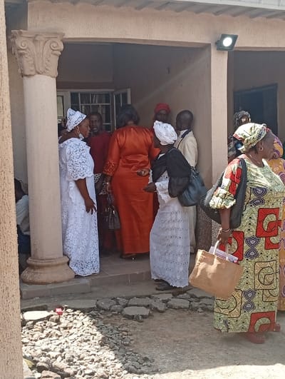 The image depicts a group of approximately ten individuals gathered at the entrance of a building in Unguwar Gaya, Nigeria, under bright natural light. The building exhibits light-colored, textured walls and a prominent decorative column with a Corinthian-style capital on the left side of the frame. A modern black floodlight is affixed to the exterior wall above the right side of the recessed entrance, emitting light. The ground directly in front of the entrance is an uneven, cracked concrete or paved surface interspersed with loose gravel and debris; a small red object is visible on the ground near the column's base.
In the foreground on the right, a woman wearing a multi-colored patterned dress, primarily green, red, and yellow, with a matching headwrap, stands facing right. She carries a dark shoulder bag and a light brown canvas tote bag in her right hand. To her left and slightly behind, another woman in a white lace dress and matching headwrap wears a dark face mask. Near the column on the left, a third woman in a white lace dress with bell sleeves and a patterned white headwrap, wearing a light blue surgical mask, stands facing right, engaged in conversation. Deeper within the building's entrance, a woman in a rust-orange dress is visible from the back, facing inwards. Several other individuals, including men in a red shirt and a light-colored jacket, and additional women in traditional attire, are partially obscured in the shadows of the doorway, appearing to congregate or move through the entrance. One man within the doorway is observed wearing a red cap.