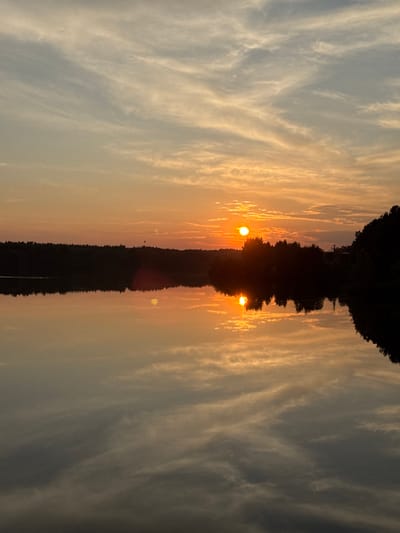 The image depicts a serene sunset over a calm body of water, likely a lake or river, in Kamysh, Russia. The sky is painted with warm hues of orange and yellow, streaked with wispy clouds that reflect beautifully on the water's surface. A silhouetted treeline borders the water, with a few faint structures visible in the distance. The sun, a bright orb, is partially obscured by trees, casting a radiant glow and a distinct reflection on the water. There are no people or animals visible in the scene, and no apparent actions or interactions taking place. The environment is tranquil and natural, characterized by the interplay of light, water, and vegetation.