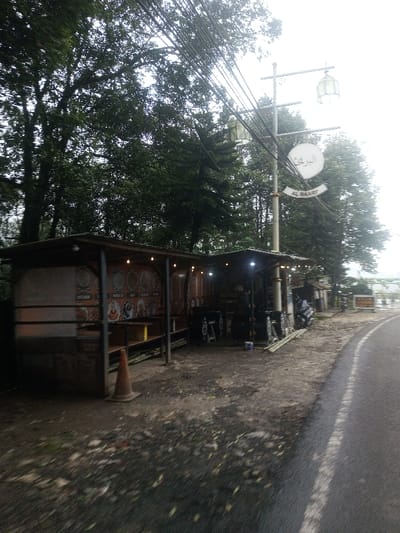 This image depicts a roadside scene in Siruppiddy, Sri Lanka, under an overcast sky. The foreground features a dirt and gravel shoulder transitioning into an asphalt road on the right, marked by a solid white line. An orange traffic cone stands on the dirt path.
Two open-fronted, rustic stalls or small shops with dark, pitched roofs are visible on the left side. The left stall displays multiple circular menu boards or decorative plates featuring images of food and drinks on its rear wall and has string lights illuminated under its roof. The right stall also exhibits illuminated string lights and appears to be a vendor setup, with a dark-colored scooter or motorcycle parked nearby.
A tall, grey metal utility pole with numerous overhead cables stands between the stalls and the road. Attached to this pole are two ornate street lamps with translucent, light-colored covers. Below these lamps, a circular white sign displays Arabic script (البركة) and "AL BAARI" in English on a curved banner.
The background is dominated by dense, dark green trees forming a thick canopy, suggesting a heavily vegetated area behind the structures. Further down the road on the right, a yellow sign is partially visible. No people are present in the scene, and no specific actions or interactions are depicted.