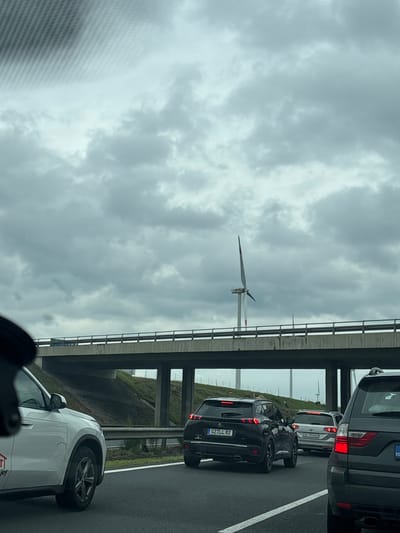 The image depicts a traffic jam on a highway, with multiple cars lined up and their brake lights illuminated. An overpass crosses above the highway. In the distance, a wind turbine stands against a cloudy sky. The location is identified as Kisigmánd, Hungary.