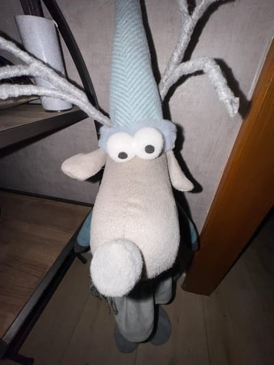 A stuffed animal rests at an oblique angle. Its head is light blue with a textured herringbone pattern and large, round white eyes with black pupils. Several grey, stringy appendages extend from its head. Its body is a solid light grey, and it has floppy ears of the same color. Its rear end has similar grey fabric with a blue accent. The animal is positioned on what appears to be a black metal rack with cylindrical objects attached. The background consists of a wooden surface and an off-white wall.