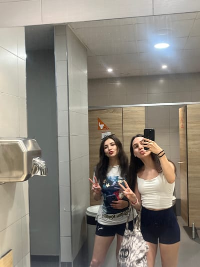 Two young women are taking a mirror selfie in what appears to be a public restroom in Palma, Spain. The woman on the left is wearing a graphic t-shirt and dark shorts, while the woman on the right is wearing a white tank top and dark shorts. Both women are posing with peace signs. In the background, there is a hand dryer, tiled walls, and wooden bathroom stall doors. The lighting suggests the photo was taken indoors, possibly in a commercial establishment or public facility.