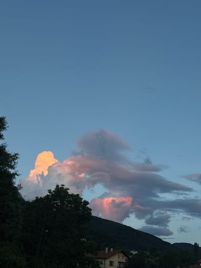 This media file depicts a twilight scene of clouds illuminated by the setting sun over a mountainous landscape. The sky is a gradient of light blue, transitioning to darker hues near the horizon. Dramatic clouds, tinged with vibrant oranges and pinks, dominate the right side of the frame, suggesting the last rays of sunlight catching their edges.

The background features a dark, tree-covered mountainside, on which a building with red-tiled roofs is partially visible. In the foreground, the silhouettes of trees are prominent, their dark foliage contrasting with the illuminated sky.

The overall mood is serene and picturesque, characteristic of a peaceful evening. The time of day is clearly sunset or twilight, given the colors of the clouds and the low angle of the sun. The location cues point to a mountainous region, possibly a village nestled in the foothills, as suggested by the building and the steep terrain.

No people or specific objects are clearly discernible, beyond the natural elements of sky, clouds, trees, mountains, and the building. There is no visible text in the media file.