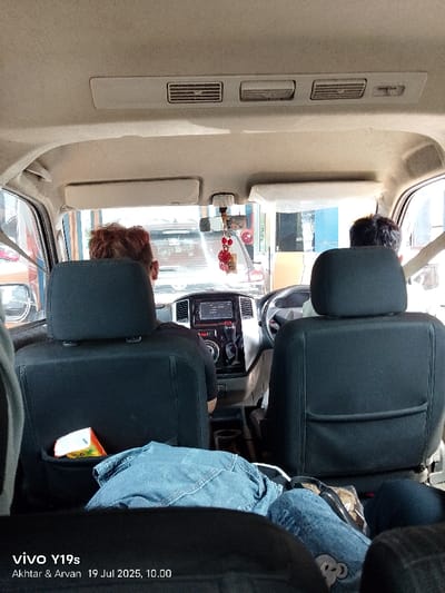 An interior view of a multi-purpose vehicle (MPV) or van, captured from a rear passenger seat, depicts the backs of two occupants in the front. The individual in the front passenger seat (left) has reddish-brown hair and wears a dark shirt. The driver (right) has dark hair. The vehicle's dashboard includes a central infotainment system and climate controls. A red ornamental tassel hangs from the rearview mirror. Rear air conditioning vents are visible on the vehicle's ceiling. A white and orange box is partially visible within the seat-back pocket of the front passenger seat. A folded light-blue denim jacket or jeans rests on the lap of a person in the immediate foreground, with a portion of a white object beneath it. Through the front windshield, other vehicles are visible, along with parts of a large structure or building featuring blue and orange elements, suggestive of an urban setting or a toll gate. The scene is situated within the Special Capital Region of Jakarta, Indonesia. The image metadata indicates it was captured on 19 Jul 2025 at 10:00.
