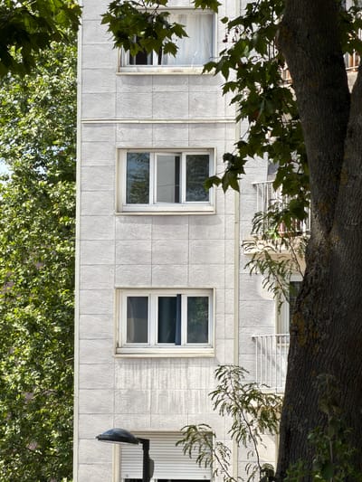 This image captures an upside-down view of a building facade and surrounding foliage. The building is clad in light-colored, textured tiles, with three visible windows. The windows are rectangular with white frames. The left-most window appears to be covered by a translucent material, possibly plastic sheeting. The other two windows show reflections of the sky and possibly some interior elements. Above the windows, a balcony railing is visible, suggesting the building has multiple stories.
Overhanging the building from the top of the frame is a thick tree branch with rough bark. Green leaves from this tree partially obscure the upper portion of the building. Below the building, a dense canopy of green leaves from another tree fills the bottom of the frame, creating a stark contrast with the building's architecture. The overall impression is one of nature encroaching on a man-made structure.
The lighting suggests it is daytime, with sunlight casting shadows and highlighting the textures of the building and tree bark. The weather appears to be fair, with a clear sky implied by the reflections in the windows. There is no visible text in the image. The scene is likely a residential or commercial building in an urban or suburban setting in Évry-Courcouronnes, France. The upside-down perspective adds an unusual and somewhat disorienting element to the composition.