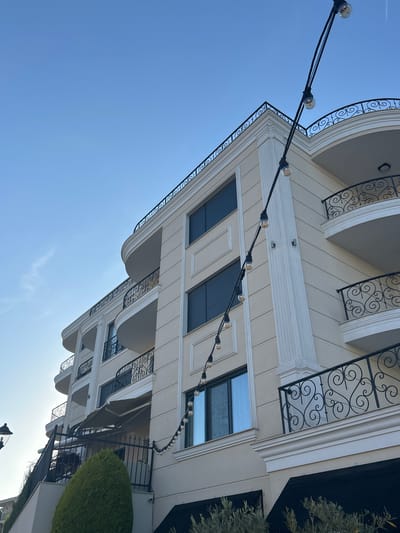 The image shows a modern building with cream-colored exterior walls and ornate black railings on balconies. String lights with clear bulbs are draped across the facade. The building features multiple floors with dark-framed windows and decorative architectural elements like columns and curved overhangs. A lush green bush is visible in the foreground on the right, and a hint of a tree can be seen behind it. The sky is a clear, bright blue with a few wispy clouds, suggesting a sunny day. The angle of the photograph is looking upwards towards the building, with the sky occupying a significant portion of the frame. The overall impression is one of a well-maintained, possibly residential or commercial building, set in an outdoor environment with pleasant weather. The scene does not depict any people or specific activities. The time of day appears to be daytime due to the bright sunlight and clear sky. The location context provided is Chernomorets, Bulgaria.