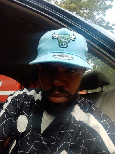 The image shows a close-up selfie taken from inside a vehicle. The main subject is a man wearing a light blue baseball cap with a green bull logo and a black and white patterned shirt. He has a beard and appears to be looking towards the camera. In the background, another person is visible in the back seat, their face obscured. The interior of the car, including the headrest of a seat, is also visible. Outside the car, trees and the sky can be seen through the window. The lighting suggests it is daytime, possibly overcast due to the diffuse light. There is some text visible on the brim of the cap, which appears to be a brand label. The image provides a candid glimpse of a moment captured while traveling in Iju, Nigeria.