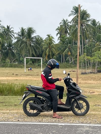 The image depicts a person sitting on a black scooter, wearing a red and black jacket and a helmet. The person appears to be looking at their phone. In the background, there is a soccer goal and a young person in a blue shirt and shorts exercising on a grassy field. The scene is set against a backdrop of numerous palm trees under a cloudy sky. The location is Glokaron, Indonesia.