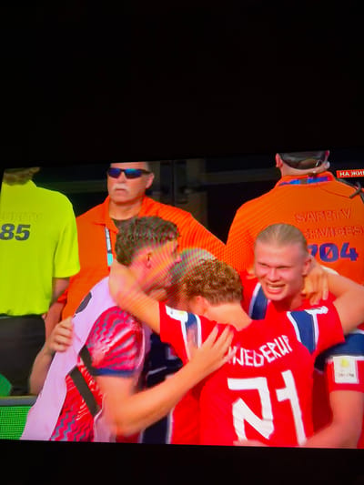 In the city of Ognianovo, Bulgaria, a group of men are engaged in a celebratory embrace on a screen. The men are wearing athletic jerseys, one predominantly red with white lettering and numbering, and another a pinkish hue with red and black wavy patterns. Two men in orange vests, labeled "SAFETY SERVICES" and displaying the number "204" and "HA ЖИВЕ", are visible in the background, one wearing sunglasses. Another person in a lime green shirt with the number "85" is also present. The scene appears to be captured from a distance, possibly on a television or monitor.