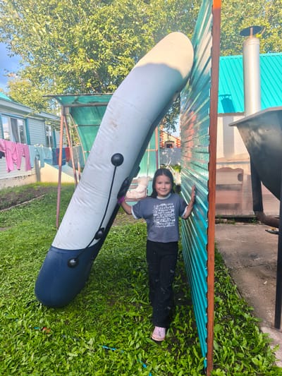 A young girl stands in a grassy yard in Burayevo, Russia, next to a large, deflated inflatable boat. She is wearing a gray t-shirt and black pants. The boat is propped against a teal corrugated metal fence. In the background, laundry hangs on a clothesline, and a house with blue siding is visible. The scene appears to be a residential area.