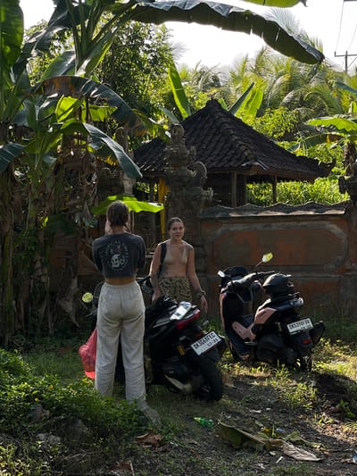 Two individuals, identified as females, are standing next to two scooters in what appears to be a rural area of Kerambitan, Indonesia. The individual on the left is wearing a dark long-sleeved crop top with graphics and light-colored linen pants. The individual on the right is wearing a light-colored bikini top and patterned shorts. Both are facing away from the camera, with the individual on the left appearing to be looking at something to their left, and the individual on the right looking towards the camera.
The environment features lush green vegetation, including large banana leaves dominating the left side of the frame. In the background, a traditional Balinese structure with a dark tiled roof is visible, suggesting a temple or a residential compound. The ground is covered in grass, dirt, and fallen leaves. One scooter is dark colored with a red accent, and the other is light pink. Both scooters have license plates. The scooter on the right has a plate that reads "DK5398FDV 08-2G". The scooter on the left has a plate that reads "DK3668FEC 03.31". A red plastic bag is visible near the legs of the individual on the left. The lighting suggests it is daytime, possibly late afternoon, with sunlight filtering through the leaves, creating shadows.