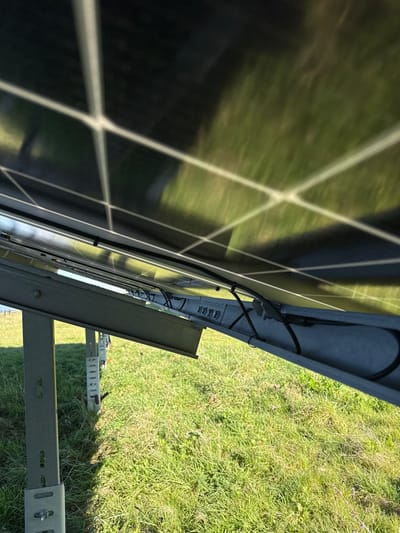 This image captures a ground-level perspective from underneath a section of a photovoltaic (PV) solar panel array in Southampton, United Kingdom. The upper portion of the frame is dominated by the dark, reflective underside of multiple solar panels, displaying their internal grid structure and reflecting the green grass below. Beneath the panels, a robust, gray metallic mounting system is visible, featuring angled support beams connected to vertical posts anchored in the ground. Bolts and fasteners are observable on the structural components. Black electrical cables are routed neatly along the underside of the panels and mounting framework, secured by black cable ties or clips. The ground visible beneath and around the installation is a sunlit field of green grass. No individuals or animals are present in the scene.