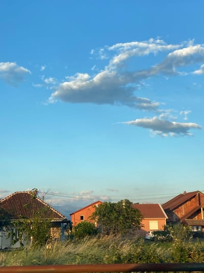 The image shows a view in Goričani, Montenegro, featuring a clear blue sky with scattered white clouds. The right side of the image includes residential houses with tiled roofs, surrounded by trees and shrubs. Overhead power lines are visible between the buildings. A vertical brown pole or structure is partially visible on the far right.