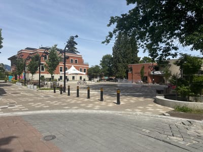 This daytime image captures a vibrant public plaza in Port Coquitlam, Canada, under a clear, bright blue sky. The scene is dominated by patterned paving stones in shades of grey, with a section of darker, red-toned pavers in the immediate foreground, where some are visibly uneven or damaged.

In the middle of the plaza, a ground-level fountain with active water jets is visible. A line of black bollards with yellow stripes demarcates an edge of the paved area. To the left, a multi-story building with reddish-orange brickwork and white trim features numerous windows and balconies, framed by young, leafy trees and modern black streetlights. A white canopy tent, displaying the text "PORT COQUITLAM" in green lettering, is set up in front of this building. Further left, a section of a colorful mural can be seen on a lower wall.

To the right, a building constructed of lighter brick and stucco stands, partially obscured by dense green trees, including a tall coniferous tree. Prominently displayed on its facade is the text "GATHERING PLACE at Leigh Square." The plaza is furnished with benches and picnic tables in shaded areas, suggesting it's designed for community gatherings and relaxation. The overall atmosphere is calm and inviting, showcasing a well-maintained urban space ready for public use. No people are visible, but the presence of the tent hints at potential events.