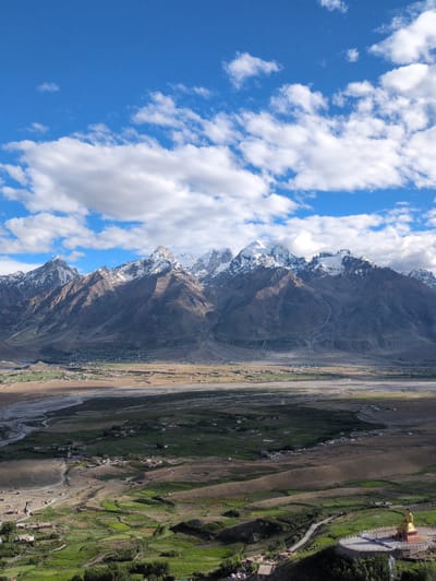 This is a vertical, high-angle, outdoor shot of the Yulang valley in Ladakh, India. The scene is dominated by a dramatic mountain range in the background, its peaks capped with snow and the slopes a mix of brown and grey. Below the mountains, the Yulang valley unfolds, a patchwork of green fields and brown, arid land bisected by a winding river. Small clusters of buildings, likely a village, are scattered across the valley floor. In the foreground, a golden Buddha statue sits atop a circular platform, a prominent landmark against the lush green slopes. The sky above is a vibrant blue, dotted with fluffy white clouds. The lighting suggests it is daytime, with shadows indicating the sun is to the left.