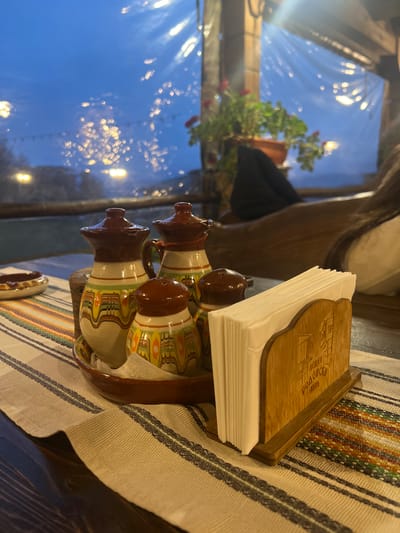 The image shows a table setting in what appears to be a restaurant or cafe in Leshten, Bulgaria.
The table is covered with a striped woven cloth. On the table, there's a napkin holder containing a stack of white napkins, and a set of salt and pepper shakers with a traditional Bulgarian design. A glimpse of a potted plant and blurred lights is visible through the window in the background.