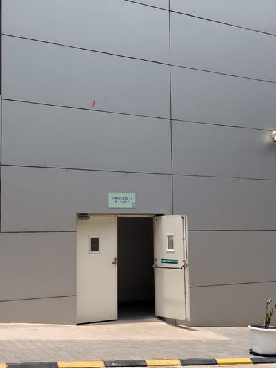 The image shows an exterior view of a modern building with a gray, paneled facade. Two outward-opening doors are visible, with one ajar. Above the doors, a light blue sign reads "STAIRCASE - 3 B-1 to B-3". To the right of the doors, a white security camera is mounted on the building. On the ground, a sidewalk with a yellow and black striped curb runs in front of the building. A white planter with a small green plant is situated near the right side of the image. The scene appears to be in an outdoor urban environment.