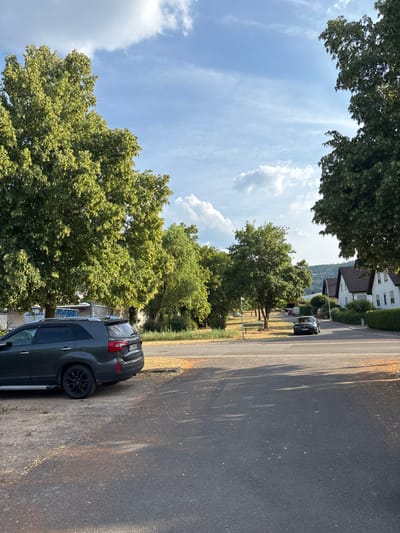 The image depicts a residential street in Bürgstadt, Germany, on a clear, cloudy day. In the foreground, a dark gray SUV is parked on a gravelly shoulder to the left. The vehicle's rear is facing the viewer, with its license plate visible. The street itself is paved asphalt, with a lane of traffic visible moving away from the viewer towards houses on the right side of the image. These houses are white with dark-tiled roofs and are surrounded by manicured hedges. Large, leafy trees line both sides of the street, casting shadows on the road. The sky is blue with scattered white clouds. The overall impression is of a quiet, suburban neighborhood bathed in sunlight.