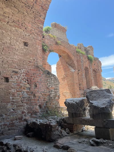This image depicts a section of the ancient Roman theater in Taormina, Italy. The foreground features rubble and architectural fragments, including large carved stone capitals resting on a stone beam. Behind these elements, a massive brick wall with evidence of disrepair and weathering forms the left side of the frame.

Dominating the midground and background is a large arched structure constructed of brick, with several smaller arched openings visible within its facade. Overgrown vegetation sprouts from cracks in the ancient brickwork, a testament to the passage of time. The sky above is a clear, bright blue. The ruins suggest a historical site that has been exposed to the elements for centuries. The overall impression is one of ancient grandeur weathered by time.