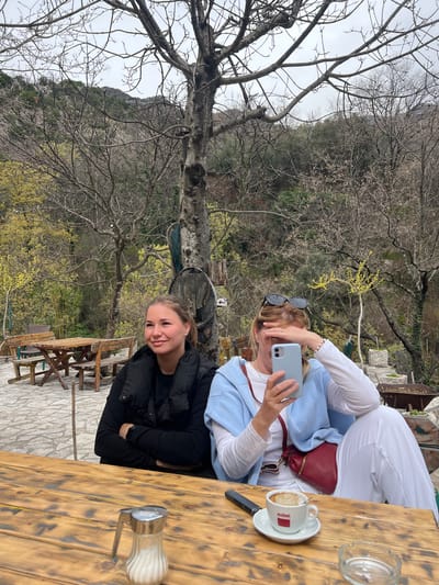 Two women are seated at a wooden table in an outdoor setting, possibly a cafe or restaurant. One woman is holding a smartphone up to her face, seemingly taking a photo. She is wearing a white top, light blue jacket, and white pants, with a small red purse. The other woman is wearing a black jacket and is smiling. A cup of coffee is on the table, along with a sugar dispenser and an ashtray. The background features a landscape with bare trees, a mountain, and greenery. The floor is tiled with stone. The location is unknown.