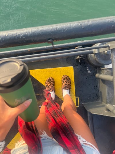 An overhead, high-angle shot shows a person's legs and a hand holding a green tumbler with a black lid on what appears to be a ferry deck. The person is wearing leopard-print clogs with white socks, ripped denim shorts, and a red and black plaid shirt loosely draped over their lap. The deck itself is dark grey, with yellow markings visible on the left side. A dark metal railing runs horizontally across the upper portion of the image, with green water visible beyond it. The person's hand, with long, light-colored fingernails, is gripping the tumbler. The overall scene suggests a casual outing on a body of water.