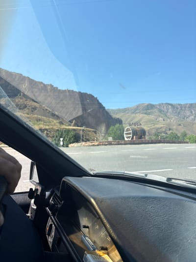 The image is taken from the interior of a vehicle, looking forward through the windshield. The foreground shows the dashboard and a portion of the driver's side door panel, with a hand visible on the steering wheel. Outside the vehicle, a paved road with white lane markings is visible, leading towards a landscape of dry, rocky hills under a clear blue sky. In the mid-ground, a large wooden barrel structure with the Armenian word "Արենի" (Areni) written on it stands on a raised platform. This landmark is located in or near the city of Areni, Armenia, as indicated by the text on the barrel. Trees and some low walls are also present in the mid-ground. The overall scene depicts a roadside view in a mountainous, arid region.