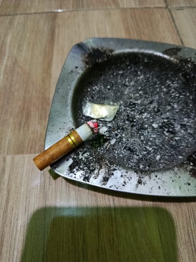 This image displays a used, square-shaped metal ashtray positioned on a light brown wooden or wood-effect tiled floor in Ketowan, Indonesia. The ashtray is heavily filled with dark gray and black cigarette ash, along with burnt cigarette remnants and a small, crumpled piece of silver-colored foil. A single cigarette, featuring a white filter with two gold bands and a brown tobacco section, rests on the left rim of the ashtray. The cigarette's tip is actively burning, indicated by a visible red ember and a small amount of formed ash. A distinct, dark shadow is cast onto the lower left portion of the wooden floor. No individuals are present or visible within the scene.