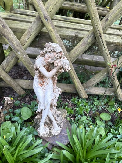 The image displays an outdoor garden scene in Stiperstones, United Kingdom, centered on a weathered light-grey statue of a female figure. The statue, depicting a young woman or maiden, is positioned leaning slightly forward, with her head resting on her bent right arm. Her left arm appears to be holding a cluster of floral elements or draped cloth against her chest. Her legs are crossed at the ankles, and she is barefoot. Her hair is long and adorned with a detailed floral wreath. The statue exhibits extensive reddish-brown discoloration and staining, particularly on the torso, arms, and hair, suggesting age and environmental exposure. It stands on an irregular, dark grey stone base, partially covered by moss.
The immediate foreground and ground around the statue consist of dark soil, interspersed with various green plants. To the left, there are clumps of broad-leaved green foliage. To the right, slender, blade-like green plants are visible, along with smaller green ground cover and scattered small yellow flowers. A thin, reddish stem with small red leaves, possibly from a shrub, extends upwards on the right side.
In the background, a rustic wooden trellis or fence is prominent, constructed from aged wooden logs or branches arranged in a diagonal, criss-cross pattern. The wood shows signs of weathering, including patches of green lichen or moss and visible fasteners. The overall scene is illuminated by natural, soft daylight.