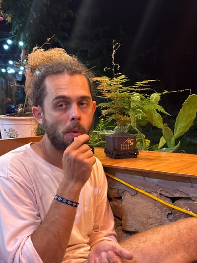 A man with curly hair and a beard is looking at the camera with his hand on his chin. He is wearing a light pink t-shirt and a blue bracelet with white diamond patterns. Behind him is a wooden structure with stone on the side, a cactus plant, and other greenery. In the dark background, there are blurred lights. The man is presumably in Mèo Vạc Commune, Vietnam.