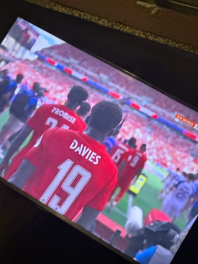 The image shows a television screen displaying a football match. On the screen, several players in red jerseys are visible on a green field. One player in the foreground is wearing a red jersey with the name "DAVIES" and the number "19" on the back. Another player behind him has "PROMISE" and a number on their jersey. In the background, there are spectators in a stadium. A banner on the screen reads "НА ЖИВО," which means "LIVE" in Bulgarian. While the location of the stadium is unknown, the presence of Bulgarian text on the banner suggests the match might be broadcast in or related to Bulgaria. The image is taken from an angle, indicating it's a photograph of a TV screen.