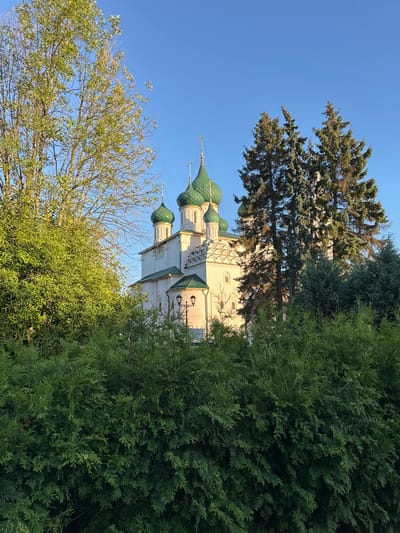 The image shows a white church with green domes and a cross on top, located in the city of Yazvitsevo, Russia. The church is surrounded by lush green trees and bushes. The sky is clear and blue, suggesting a sunny day. There are no people visible in the image. The scene appears peaceful and serene, with the natural elements complementing the architectural beauty of the church.