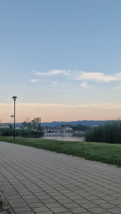 The image captures a waterside scene in Keszthely, Hungary, during what appears to be late afternoon or early evening, given the soft, warm light bathing the sky. The sky transitions from a clear, light blue on the left to a soft, peachy hue on the right, with wispy white clouds visible. A row of buildings with white facades and dark roofs lines the waterfront, reflecting in the calm water. Beyond the buildings, a range of blue-tinged hills forms the backdrop.
The foreground is dominated by a paved walkway constructed of grey rectangular tiles, laid in a diagonal pattern that recedes into the distance. To the left of the walkway is a strip of vibrant green grass, bordering the water. A dark-colored lamppost stands on the walkway, its light fixture pointing downwards. A small white sign with blue text, possibly indicating directions or facilities, is attached to the lamppost. The overall atmosphere is peaceful and serene, suggesting a leisurely setting. There is no discernible activity or people visible in the frame. The image is oriented sideways, with the sky appearing at the top and the walkway at the bottom of the frame.