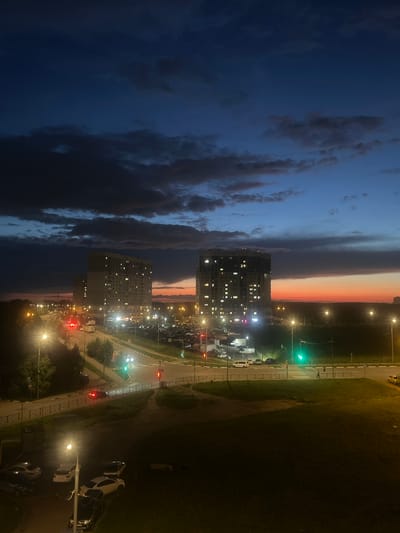 The image depicts a twilight scene over a residential area in Domodedovo, Russia. The sky transitions from a deep blue on the left, filled with dramatic, dark clouds, to a warm gradient of orange and pink hues near the horizon on the right, suggesting the end of the day. Two tall, modern apartment buildings with illuminated windows are prominent features.
Below the buildings, a street is visible, lined with streetlights casting a yellow glow. Several cars are parked along the street and in a designated parking lot. Traffic lights are also visible, one showing green and others red. The scene includes patches of grass and trees, indicating an urban or suburban parkland. The overall mood is peaceful, with the artificial lights of the city contrasting with the fading natural light of the sky. The orientation of the image is vertical. No visible text is present in the image.