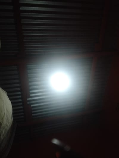 A ceiling is constructed of corrugated metal sheets supported by reddish-brown beams. A bright, diffused light source dominates the center, obscuring the details of the metal. To the left, a soft, white object, possibly fabric, is visible. The rest of the scene is shrouded in darkness, making it hard to identify. At the bottom, some blurry light spots can be seen.