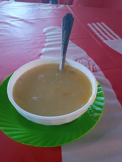 A close-up image captures a bowl of soup or porridge set on a table, likely in a casual eatery in Namanga, Kenya. The main subject is a white, round bowl filled with a thick, light beige or creamy yellow liquid. Small bubbles are visible on the surface, and tiny, light-colored flecks, possibly indicating vegetables or grains, are suspended within the liquid. A metallic spoon, featuring an ornate, embossed design on its handle, is placed upright in the center of the bowl.
The bowl rests on a bright green plastic plate or saucer, which has a distinct leaf-like design with prominent ridges. This vibrant green contrasts sharply with the other elements. Beneath the plate is a red and white checkered tablecloth. A significant detail on the tablecloth is the visible text and design of the "Coca-Cola" logo. The word "Cola" is clearly discernible in its iconic script, along with parts of the "Coca" portion of the logo.
The scene appears to be set during the day, indicated by the bright, natural lighting. The arrangement suggests a meal about to be consumed. No individuals are present in the frame, keeping the focus entirely on the food and its immediate surroundings. The overall setting implies a local, unpretentious dining experience, common in such regions.