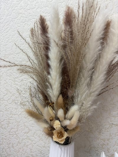 The image displays a detailed close-up of a decorative arrangement primarily composed of dried natural elements, set against a textured white wall. The arrangement features a variety of dried grasses and flowers, presented in a fan-like or cascading style.
The most prominent components are large, fluffy plumes, likely pampas grass, which range in color from creamy white to light beige and a deeper, muted brown or purplish-brown. These plumes have a soft, feathery texture and are arranged to spread out towards the left side of the frame. Mixed in with these larger elements are finer, wispy dried grasses with delicate seed heads, adding subtle texture and depth. These finer grasses are also in neutral shades of light brown and beige.
Towards the right side of the arrangement, a cluster of smaller, softer, and more rounded fluffy elements, possibly dried bunny tail grass (Lagurus ovatus), is visible in shades of light beige and warm brown. Within this cluster are a few dried floral accents: a light cream or pale yellow flower with a distinct, slightly darker brown center, and a smaller, cream-colored, fluffy dried flower next to it. Additionally, a few dark, spherical seed pods or berries on thin stems add a touch of darker contrast.
The arrangement is held within a white, ribbed or fluted vase, a section of which is visible on the far right edge of the image. The background is a white or off-white wall with a visible, rough stucco or textured wallpaper pattern, providing a subtle, complementary backdrop. The lighting is soft and even, emphasizing the varied textures and natural color palette of the dried botanicals. The overall aesthetic is natural, rustic, and decorative. The image appears to be rotated 90 degrees clockwise from its likely upright display orientation.