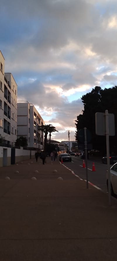 This photograph captures an urban street scene in Aïn El Aouda, Morocco, during the late afternoon or early evening. The sky is partially cloudy, with patches of blue visible and the setting sun illuminating the clouds with soft oranges, pinks, and warm grays.
On the left, several multi-story modern residential buildings with white or light-colored facades and balconies line the street. Their windows reflect the subdued light. A wide pedestrian sidewalk, dotted with concrete spherical bollards, runs along the base of these buildings. A few people are walking on this sidewalk, their figures largely silhouetted against the ambient light.
In the midground, a paved road stretches into the distance, with several cars either parked or in motion. A black SUV is prominently visible on the road. Orange traffic cones are placed along the side of the road, possibly indicating a temporary obstruction or work area. Several tall palm trees are visible between the buildings and the road, adding a distinct tropical touch. Further down the street, a metallic utility structure, possibly an antenna or cell tower, can be seen.
On the far right, a dense canopy of dark green trees forms a stark contrast against the brighter sky, casting long shadows. Two street signs or posts, one with a circular blue sign and another with a rectangular white sign, are present on the right edge of the frame. The overall atmosphere is tranquil, marking the transition from day to dusk in a bustling urban environment. No legible text is visible in the image.