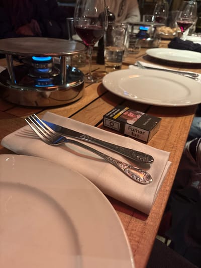 The image depicts a dining table scene in a restaurant in La Clusaz, France. The foreground features a partial view of a white ceramic plate. To its right, a cream-colored cloth napkin is placed, holding a fork and a knife with ornate handles. Adjacent to the napkin, a black pack of Marlboro Red cigarettes lies on the wooden table, displaying the French health warning "Fumer tue" (Smoking kills) and a graphic image.

In the mid-ground, a stainless steel fondue burner with a blue flame is active, positioned beneath a flat metal surface. Behind this, a wine glass filled with red wine and a clear glass of water are visible. Further back, another white ceramic plate is set, accompanied by a fork and another wine glass containing red wine. The background is blurred but shows indistinct figures of people seated at the table, suggesting a social dining environment. The table surface is constructed from light-colored wood planks.