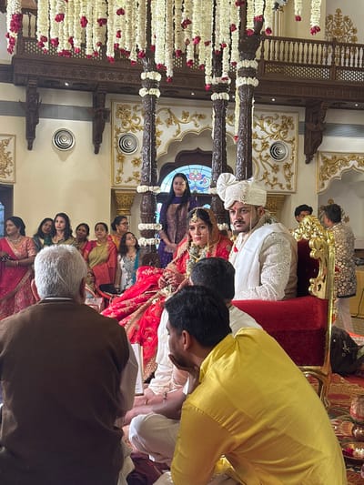 An indoor Hindu wedding ceremony is depicted in a grand hall with traditional Indian architecture, located in Vadodara, India, likely within a palace or heritage venue. The scene is dominated by a central mandap structure featuring four ornate, dark wooden pillars extensively draped with strings of white and red flowers. Hanging floral garlands also descend from the high ceiling, which exhibits intricate wooden beams and carvings.
At the center, the groom is seated on a red velvet chair, wearing a cream-colored sherwani with embroidery, a matching turban, and a pearl necklace. He has a mustache and a focused expression. To his left, the bride sits on a similar chair, adorned in a red lehenga-choli with gold embroidery, elaborate jewelry including a mang tikka and nose ring, and a red veil partially covering her head. Her hands are clasped as if participating in a ritual.
In the immediate foreground, two men are seated on a red carpet with their backs to the camera, facing the couple. The man on the left has grey hair and wears a brown waistcoat over a white shirt. The man on the right wears a yellow kurta and is seen with his right hand near his chin, attentive.
Behind the bride and groom, a woman in a dark purple embroidered outfit stands observing. To the far left, a group of approximately eight women in various traditional Indian attire (sarees, salwar kameez) stand and observe the ceremony. Additional guests are indistinctly visible in the background.
The walls are light-colored with intricate gold and white plasterwork, archways, and traditional motifs. Circular ventilation grilles are visible on the wall. A decorative wooden balcony with intricate railing is present in the upper background, above a large arched window with a stained-glass design. Small metallic ritual objects are visible on the floor in front of the seated men. The lighting is bright, illuminating the festive and formal environment.