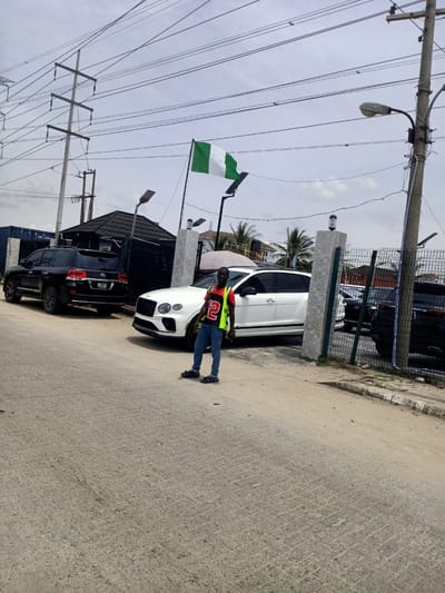 A daylight, eye-level outdoor shot captures a street scene in Eputu Town, Nigeria. A man stands on a light-colored paved road, facing forward, with multiple vehicles parked behind him. He wears a red and black t-shirt featuring a large white number "2", a yellow high-visibility vest, blue jeans, and dark sandals.

Behind the man, a black Toyota Land Cruiser SUV is parked on the left, displaying a license plate that reads "LAGOS STATE JJK 199FH". To its right, a white Bentley Bentayga SUV is partially visible. Further right, behind a dark metal mesh fence with concrete posts, other dark-colored cars are parked. A small building with a dark roof is situated behind the black SUV.

Above the vehicles, a Nigerian flag (green, white, green vertical stripes) flies on a pole. The sky is overcast, and numerous power lines traverse the upper portion of the frame, supported by utility poles. Several streetlights are also visible, some equipped with small solar panels. On the far left, a blue shipping container is partly visible, bearing markings such as "CTNU 328089 9". Palm trees can be seen in the background on the right side of the image, hinting at a tropical or subtropical setting. The overall scene appears to be a commercial or residential area with an emphasis on vehicles.