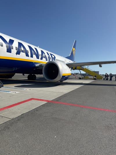 The image shows the side of a Ryanair airplane parked on an airport tarmac. The airplane is predominantly white with blue and yellow accents. The word "RYANAIR" is prominently displayed in large blue letters along the fuselage. The engine of the plane is visible, as is part of the wing with the Ryanair logo. The plane is positioned on a concrete surface marked with red lines, indicating it is on a taxiway or apron.
In the background, a yellow baggage loader or passenger stairs vehicle is connected to the aircraft. Several people are visible near the vehicle, appearing to be ground crew or passengers. Another aircraft is partially visible further in the distance. The sky is a clear, bright blue, suggesting a sunny day with good weather. The sun appears to be high in the sky, indicating it is likely daytime, possibly late morning or early afternoon.
The scene is set at an airport, likely Kastrup, Denmark, as indicated by the location context. The overall mood is functional and operational, typical of an airport environment. There is no discernible emotion evident in the image. The visible text includes "RYANAIR" on the plane and some text on the yellow vehicle, but it is too small and blurry to be read clearly.