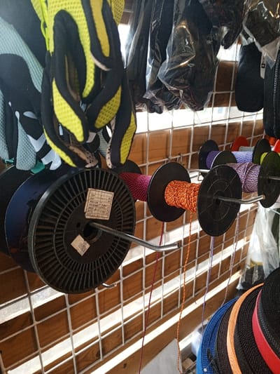 The image captures a detailed close-up of retail merchandise displayed on a white grid-patterned metal wall rack inside a shop in Baza, Myanmar. The background behind the rack consists of light-brown wooden paneling.
In the upper left quadrant, a pair of black and neon yellow mesh cycling or athletic gloves hang vertically. To their right, several other dark-colored gloves or accessories, some individually wrapped in clear plastic, are also suspended from the rack.
Across the middle of the frame, a series of black plastic spools of cord are mounted on curved metal hooks protruding horizontally from the grid. From left to right: a large, partially empty black spool with a white label; a smaller spool wound with magenta cord; a spool with bright orange cord trailing downwards; a spool with purple cord also trailing; and a partially visible spool with light blue cord. The cords are thin and braided.
In the lower right, a stack of flat, rolled webbing or straps is visible, displaying colors including dark blue, black, orange, and red. No people are visible in the scene, and no specific actions or interactions are depicted beyond the static display of merchandise.