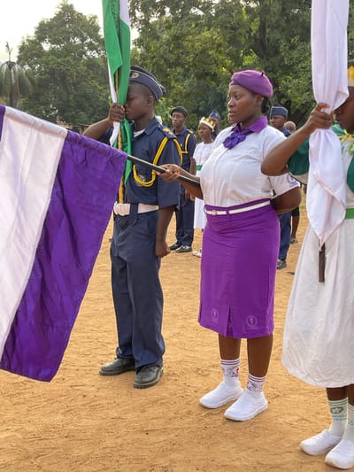 The image shows a group of individuals in Tabu, Nigeria, assembled outdoors. The ground is bare earth. In the foreground, a person in a dark blue uniform, possibly military or a similar service, holds a pole with a flag composed of white, green, and purple stripes. The person appears to be saluting the flag. Other members of a group stand behind them, some in purple skirts and white shirts, and others in white dresses with green accents. All seem to be participating in a formal event or parade. Some of the group are holding white cloths or flags.