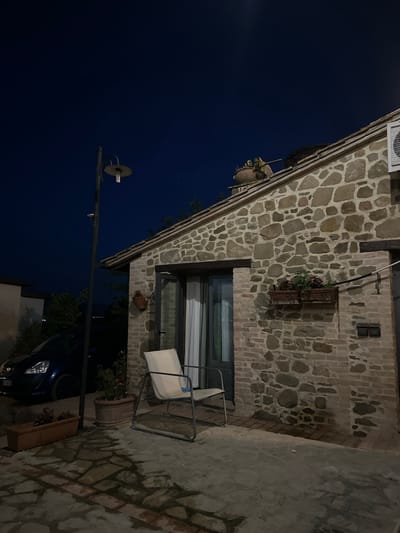 A nighttime exterior view of a stone building in Cannara, Italy, shows a single white metal chair positioned on a paved patio in front of a double door. To the left of the building, a dark blue car is parked. A black metal lamppost stands on the patio, casting light onto the scene. A planter with green plants sits near the car. The sky is dark blue, indicating nighttime. The building's facade is constructed of irregular, rough-hewn stones, with some visible brickwork at the lower section and around the doorway. A window box with plants is mounted on the stone wall above the planter.