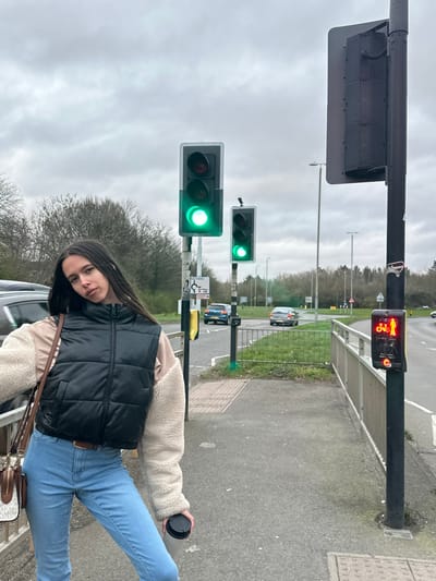 The image depicts an outdoor urban road environment under an overcast sky, specifically at the A6003/A43 Phoenix Parkway Roundabout in Corby, United Kingdom.
A female individual is centrally positioned in the foreground, standing on a paved sidewalk. She has long, dark hair parted in the middle and is looking towards the camera with her head tilted slightly. She wears a dark-colored puffer vest over a beige long-sleeved top with contrasting cream-colored fleece sleeves, light blue jeans, and a brown belt. A brown shoulder bag with a visible "MK" monogram charm hangs over her right shoulder. Her left hand holds a black travel mug.
To the individual's right, a black pole supports a pedestrian/cyclist signal light displaying a red illuminated symbol of a standing person and a bicycle, indicating "do not cross." Adjacent to this pole is a metal guardrail separating the sidewalk from the road. Further into the midground, two vertical traffic light poles are visible, both displaying green lights for vehicular traffic. Below the left traffic light, a small white sign with text and symbols is attached.
The road surface is asphalt, and a grassy median separates traffic lanes. In the background, three vehicles are visible on the road: a dark-colored SUV partially obscured on the left, a blue hatchback, and a silver sedan in the distance, all consistent with left-hand drive vehicles common in the UK. Distant road signs indicating a roundabout (red triangle with circular arrows) and route numbers (A6003, A43) are discernable. The background features bare deciduous trees, suggesting a non-summer season. No direct interactions between individuals are observed; the female individual is posing for the photograph.