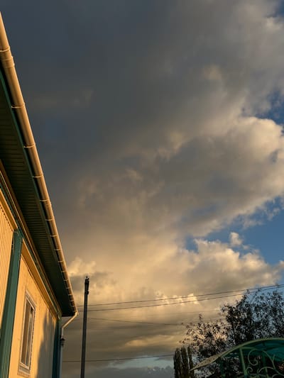 This media file depicts a dramatic sky with heavy, dark clouds gathering on the left, contrasting with lighter, sunlit clouds on the right. The scene appears to be an outdoor setting, possibly a residential area, with the roof of a building in the foreground. The building's roof is made of corrugated metal, with a light-colored gutter system. A window is visible on the side of the building, which also appears to be clad in corrugated metal.
The sky suggests changing weather, possibly a storm approaching or passing. The lighting indicates it's either early morning or late afternoon, with the sun casting a warm glow on the clouds from the right. Several utility poles and wires are visible, stretching across the frame and adding to the urban or semi-urban context. A dark tree with sparse foliage and a decorative green fence are also present on the right side of the image, further contributing to the impression of a developed area.
No people are visible in the frame. The overall mood is somewhat ominous due to the dark clouds, yet also majestic and awe-inspiring due to the play of light and shadow in the sky. The location is identified as Starobaltachevo, Russia. There is no visible text in the image.